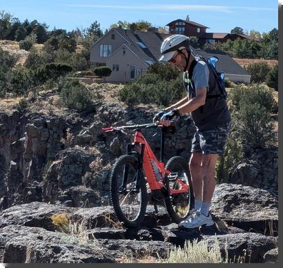[A man dressed for biking, with a white helmet, stands next to a small bright orange mountain bike, on some rocks at the edge of a cliff]