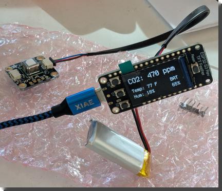 [A sensor and small microcontroller, connected by a cable, sitting on pink anti-static bubblewrap. The microcontroller is also plugged in to a USB cable and a battery; it has a built-in display that reads: CO2: 470 ppm, Temp: 77F, Hum: 18%, BAT 65%]