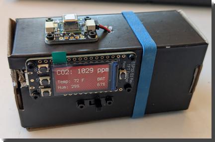 [A small black cardboard box that holds a microcontroller with a small display, reading CO2: 1029 ppm, Temp: 72F, Hum: 29%, BAT 67% all in white on a red background. On top of the box is the small sensor that's taking the CO2 readings. The box is held closed with a blue rubber band. There's a small power switch on the front under the microcontroller.]