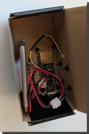 [Looking down into a small cardboard project box. There's the underside of a microcontroller, with a cable that runs from it out through a hold in the side of the box. Then there's a power cable, red and black, that runs from a power switch through a JST plug to the microcontroller and a slim li-po battery that's velcroed to the side of the box. All the components are screwed to the sides of the box with black machine screws and nuts.]