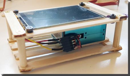 [a boxy blue air quality sensor in a crate made out of popsicle sticks, with a microcontroller with screen on top]
