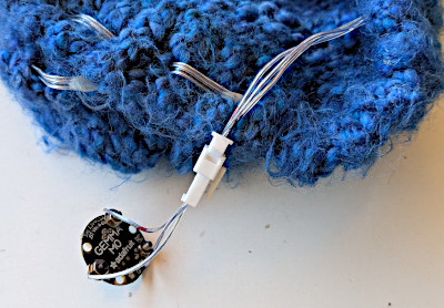 [a microcontroller the size of a qyarter, plugged in to a light string wound through a blue knit cap]