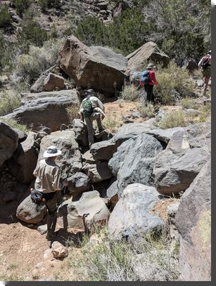 [A hiker scrambles up a 6-foot tall pile of boulders, with another hiker waiting to ascend and two others already on top]