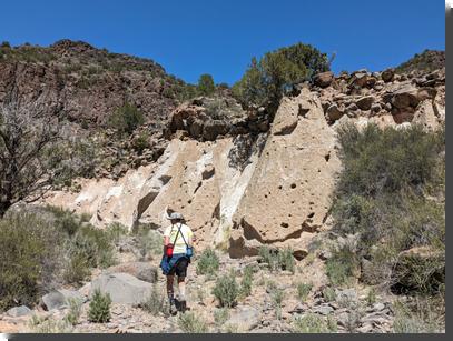 [A hiker walks next to a pair of 'tent rocks' formed of tuff. They're about 3-4 times the hiker's size.]