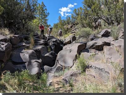 [Hikers stand at the top of a pour-off fifteen or twenty feet high, of basalt that has been worn into smooth and complex curves]