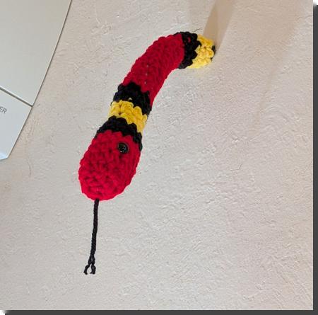 [a crochet kingshake, red with bands of black and yellow and with a black yarn tongue, protruding from a hole in a wall]