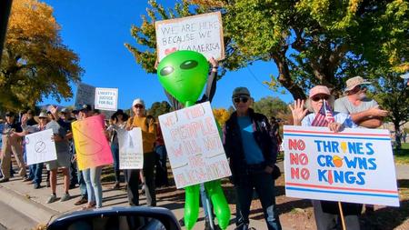 [A row of protesters standing on the edge of a sidewalk facing a street, holding a variety of signs. In the center someone is holding a large (more than human sized) inflatable alien and a sign: WE THE PEOPLE WILL NOT BE ALIENATED]