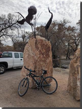 [A mountain bike leans against a large, beautiful rock atop which are metal sculptures of two cranes, one taking off and the other standing with head pointed toward the sky]