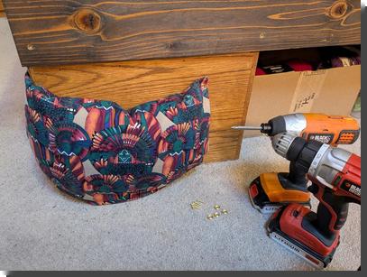 [A colorful pillow screwed around the corner of a bed frame, with a cordless drill and impact driver and a pile of brass screws and finishing washers]