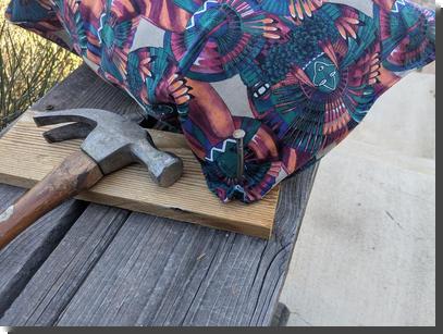 [A colorful pillow with one corner nailed to a board with a very large nail, and a hammer sitting nearby]