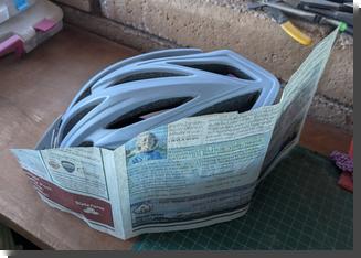 [a piece of newspaper curled around the left side of a bike helmet with the front of it cut down to match the curvature of the helmet, and the rear part curving around the back of the helmet]