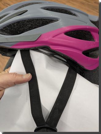 [holding a piece of paper up to the straps on a bike helmet to make a pattern for an ear-warmer that fits over the straps]