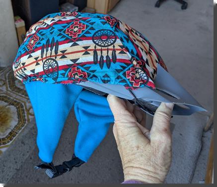 [a hand holding up a bike helmet covered with a blue and tan cloth cover, not completely finished, with fleece ear-warmers attached]